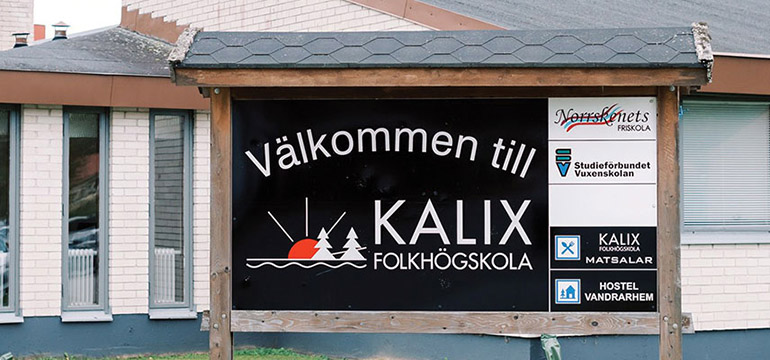 En svart skylt med texten Välkommen till KALIX folkhögskola, framför en vit byggnad